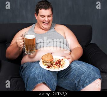 Something to wash down his meal. An obese young man sitting on a sofa and gorging himself on beer and fast food. Stock Photo