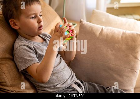 Cute baby boy assembling multicolored Rubik's cube solving logical game lying on couch at home Stock Photo