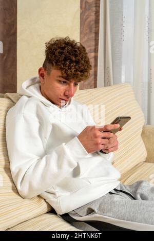 Caucasian young man with curly hair and a sporty style of clothes sitting on a white sofa typing messages on his smartphone, he is writing to his Stock Photo