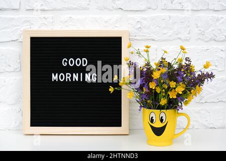 Text Good morning on black letter board and bouquet of colored flowers in yellow cup with painted funny face on table against white brick wall. Creati Stock Photo