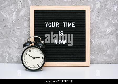 Your time is now. Motivational quote on letter board and black alarm clock on table against stone wall. Concept inspirational quote of the day. Stock Photo