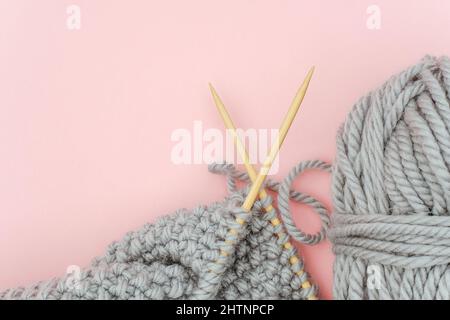 Piece of grey knitted fabric on bamboo wood needles with ball of yarn, process of knitting on pink background. Copy space for text .Top view Flat lay Stock Photo