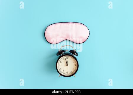 Pink sleep mask for eyes and black alarm clock on blue background It 's midnight. Top view Copy space. Concept eye protection from light for good slee Stock Photo