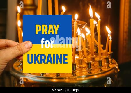 Male hand holding Ukraine flag card with Pray for Ukraine message in front of  burning candles in a church in Kiev Stock Photo
