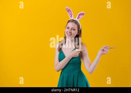 Shot happy girl in fluffy pink bunny ears, points fingers to side, demonstrates something over an empty space isolated on a yellow studio background. Easter holiday concept Stock Photo