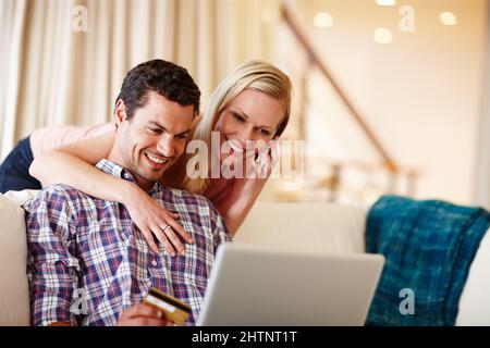 Shopping from the comfort of our couch. A beautiful woman hugging her husband from behind while he sits on the couch working on his laptop. Stock Photo