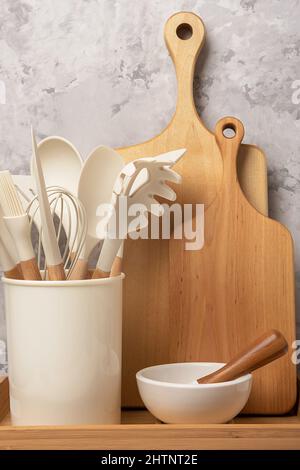 Cooking utensil set on kitchen table. Silicone kitchen tools, wooden cutting boards, mortar bowl with pestle. Front view. Stock Photo