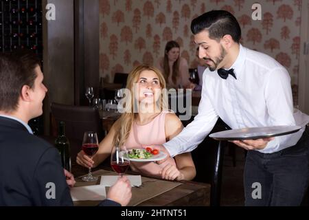 Polite waiter bringing ordered dishes to smiling couple at restaurant Stock Photo