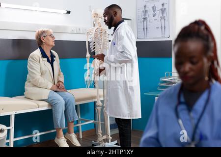 Chiropractor doctor holding anatomy human skeleton arm explaining bones pain to senior woman patient discussing medical treatment during osteoporosis appointment in hospital office. Medicine concept Stock Photo