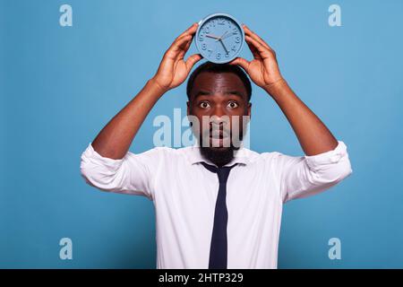 Portrait of stressed office worker holding alarm clock over head showing passing time concept on blue background. Anxious businessman in white shirt and tie looking nervous feeling deadline pressure. Stock Photo