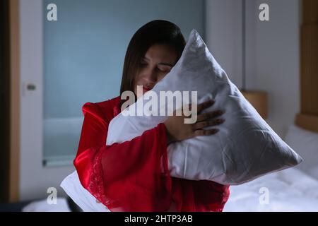 Asian woman in a silk red nightgown standing hugging pillow with two hands in a bedroom at night. Female in satin nightwear and embracing soft pillow Stock Photo