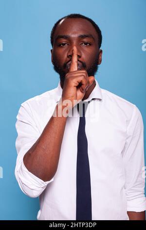 Portrait of african american man doing hush sign with index finger over lips on blue background. Calm businessman asking for silence doing shush hand gesture. Stock Photo