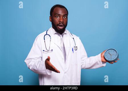 Medical doctor looking shocked running out of time holding alarm clock wearing white lab coat and stethoscope. Medic in a hurry panicking having a professional health care crisis. Stock Photo