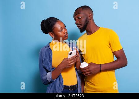 Concerned man spying on girlfriend sms on mobile phone. Young woman holding smartphone and browsing internet while her boyfriend looks at notification on screen in studio, expecting betrayal. Stock Photo