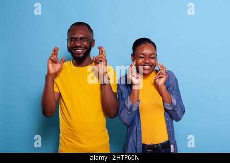 Boyfriend and girlfriend crossing fingers and hoping for good luck in studio. Joyful couple praying for miracle sign and having positive expectations, feeling superstitious about belief. Stock Photo