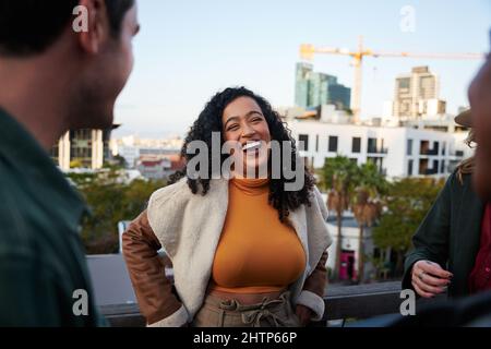 Biracial female laughing, having a good time with group of multi-cultural friends socializing on a rooftop terrace at dusk. Stock Photo