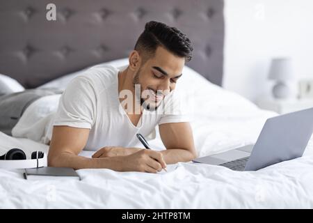 Distance Learning Concept. Millennial Arab Guy Using Laptop While Lying In Bed Stock Photo