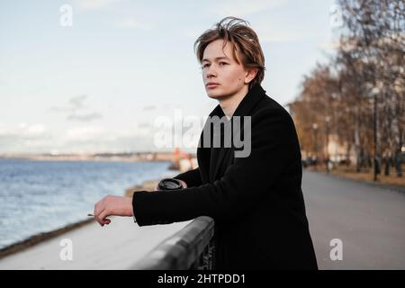 Young man in coat with cup of coffee in hands smoking a cigarette and walking on a city street in the morning. Blonde man taking a break with hot beve Stock Photo