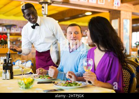 African American waiter bringing ordered dishes to smiling couple at restaurant Stock Photo