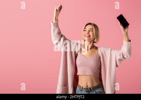 happy young woman in soft cardigan holding smartphone with blank screen isolated on pink,stock image