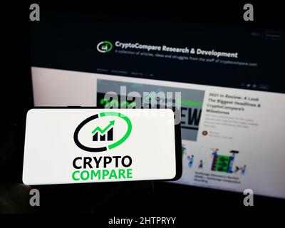 Person holding mobile phone with logo of British financial company CryptoCompare on screen in front of business web page. Focus on phone display. Stock Photo