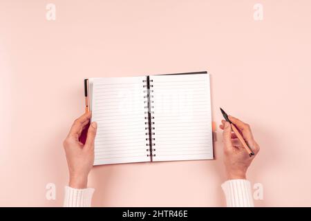 Female hand writing in blank open spiral notebook with pen on pastel pink background. Top view, copy space. Education, remote work, business, finance Stock Photo