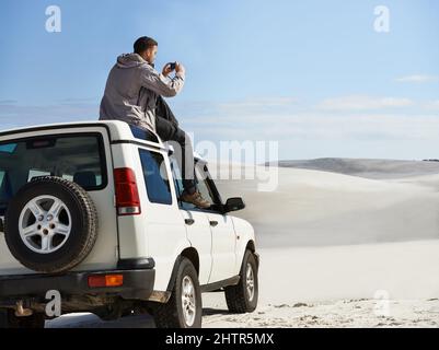 Capturing the scenery. Cropped shot of a young man sitting on his car roof while on a roadtrip. Stock Photo