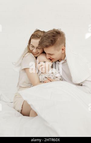 Portrait of young happy smiling beautiful family with cute cherubic infant baby in white clothes sitting on white bed. Stock Photo
