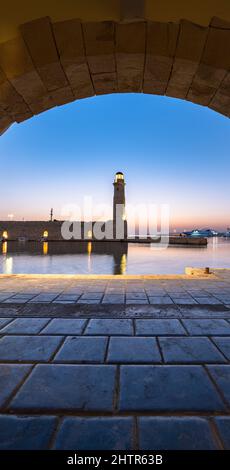Illuminated lighthouse at dusk view through a medieval arch in the old Venetian harbour, Rethymno, Crete, Greece Stock Photo