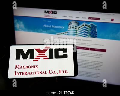 Person holding cellphone with logo of Taiwanese company Macronix International Co. Ltd. on screen in front of webpage. Focus on phone display. Stock Photo