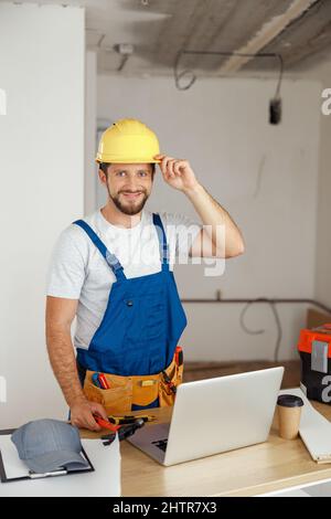 Smiling handyman worker in uniform wearing tool belt and hardhat looking at camera during renovation work indoors Stock Photo