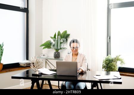 Focused and confident woman in glasses using a laptop for remote work sitting at the desk at office. Concentrated aged woman typing mails answers, messaging online Stock Photo