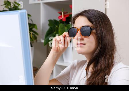 A woman looks at a computer screen through perforated glasses with holes to improve vision in astigmatism and keratoconus Stock Photo