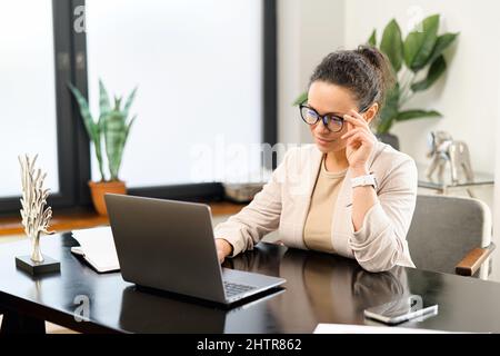 Cheerful female employee works with a laptop in the modern office. Middle-aged positive woman in smart casual wear typing on the keyboard, a journalist is writing an article Stock Photo