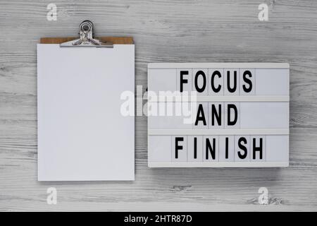 'Focus and Finish' on a lightbox, clipboard with blank sheet of paper on a white wooden background, top view. Flat lay, overhead, from above. Stock Photo