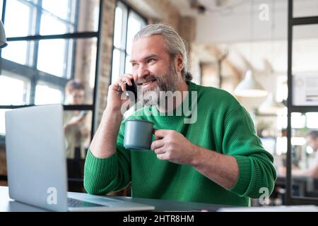 Bearded man in casual sweater drinking coffee and talking on the phone, while sitting at the table with laptop at office Stock Photo