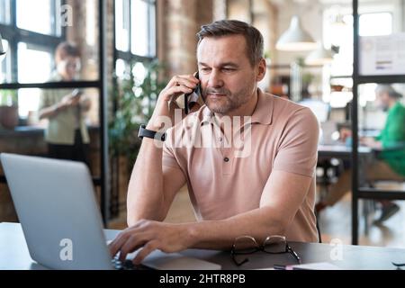 Mature businessman talking on mobile phone while typing on laptop sitting at his workplace at office Stock Photo