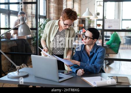 Secretary bringing the documents for signing to her boss while she working on laptop at the table at office Stock Photo