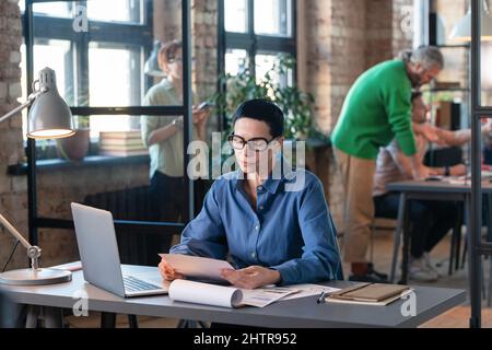 Serious businesswoman in eyeglasses reading a document sitting at her workplace in front of the laptop, she doing paperwork at office Stock Photo