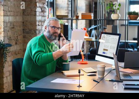 Serious bearded businessman wearing his eyeglasses to read the contract while talking on mobile phone with colleague, he working at office Stock Photo