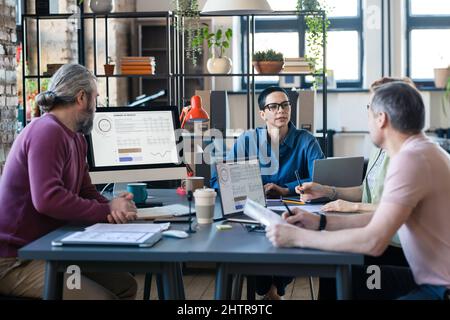 Mature businesswoman in eyeglasses sitting at the table and discussing presentation on computer monitor together with her colleagues at meeting Stock Photo