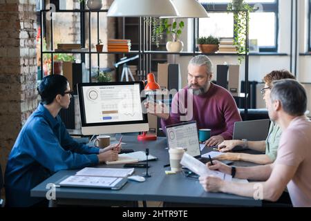 Bearded businessman pointing at computer monitor and analyzing graph on the monitor with colleagues during teamwork at meeting Stock Photo