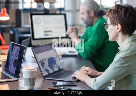 Rear view of businesswoman in eyeglasses sitting at the table and typing on laptop, she analyzing charts online with her colleagues in the background Stock Photo