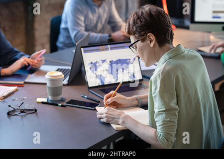 Rear view of businesswoman in eyeglasses sitting at the table with her colleagues and making notes in notebook during meeting at office Stock Photo