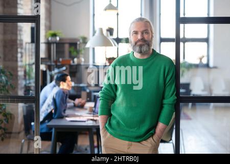 Portrait of mature bearded manager in casual clothing leaning on the glass wall and looking at camera while having meeting with colleagues Stock Photo