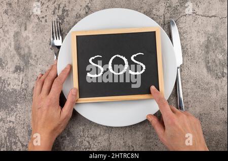 Photo of man's hands with fork, knife and an "SOS" word on a board on a plate Stock Photo