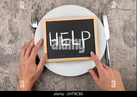 Photo of man's hands with fork, knife and a "help" word on a board on a plate Stock Photo