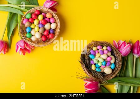 Overhead view of colorful easter eggs and candies in nests with tulips on yellow background Stock Photo