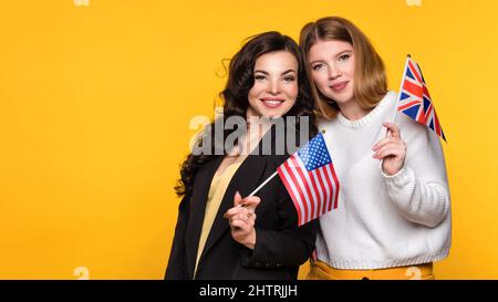 Two Girlfriends holding flags UK and USA. Study Abroad Concept. International Student Exchange Program. Young Women hug embrace and Looking at Camera on Yellow Background. Stock Photo