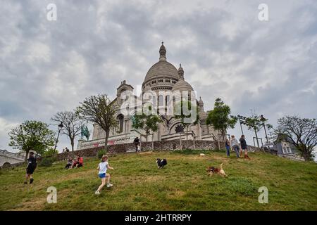 Paris, France - April 2020: during the COVID-19 lockdown, a little girl is playing with her dogs while people are resting sitting on the grass just in Stock Photo